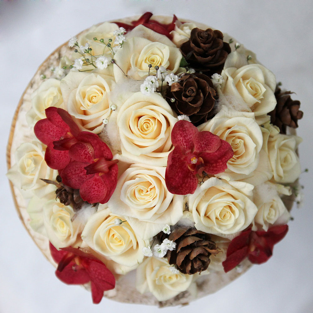 Le bouquet sur structure en écorce avec roses crème, orchidées vanda bordeaux et pommes de pins.